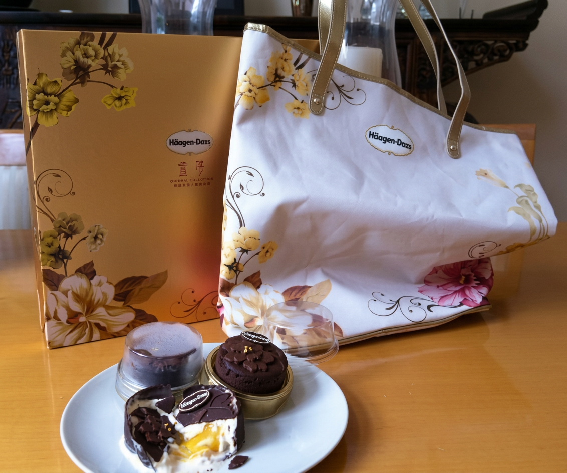 Caixa dourada da Häagen-Dazs e uma bolsa com flores, ao lado de doces gelados em um prato sobre uma mesa de madeira.
